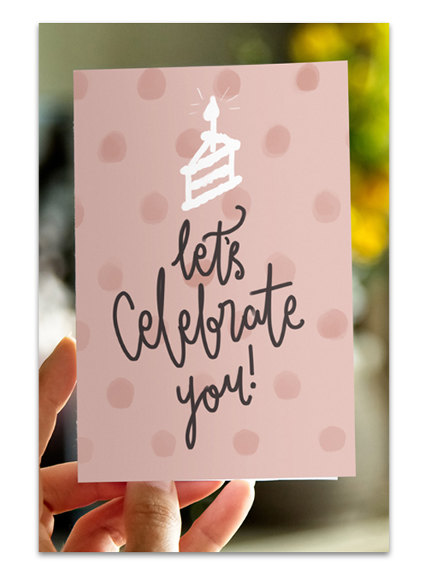 A hand holds a greeting card that features an illustration of a cake and says "Let's celebrate you!"