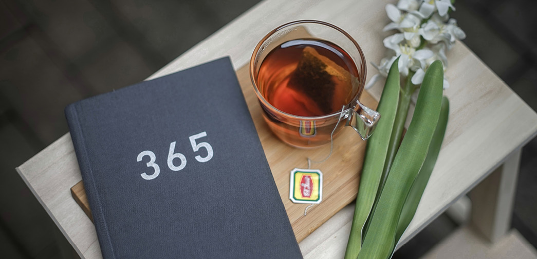 A mug of iced tea and some flowers are on a table, beside a book that reads "365" on the cover